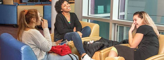 A group of three students relax in a campus lounge, smiling and laughing.